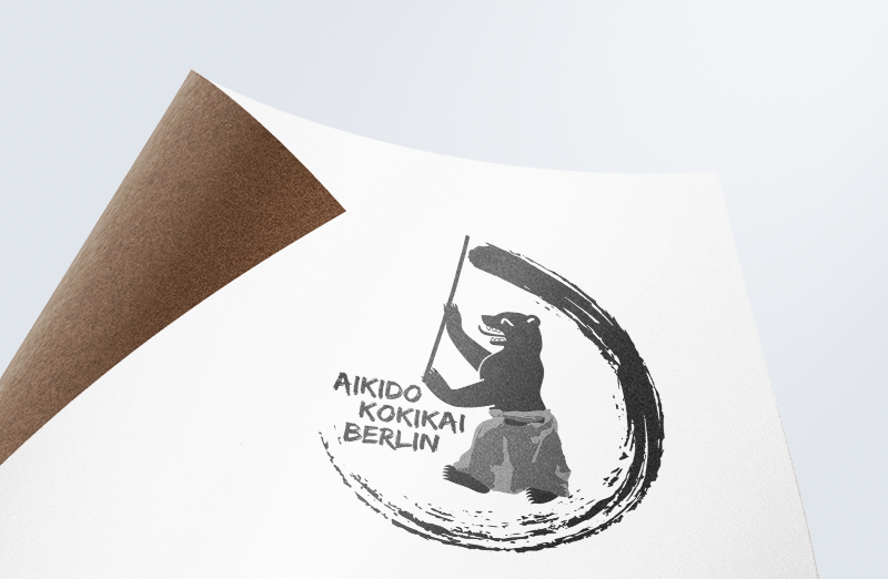 Logo auf gewelltem Papier: Tuschekreis mit Aikidoka und Stab, Schriftzug „Aikido Kokikai Berlin"